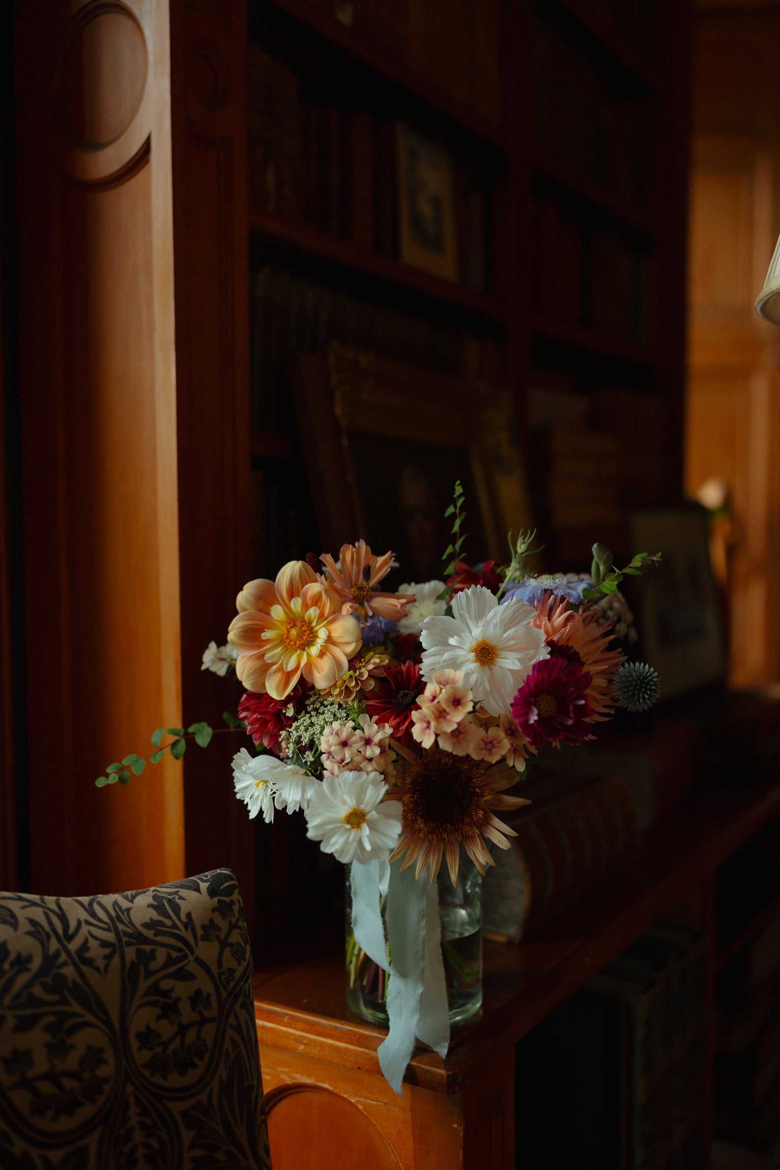A Bridal Bouquet sits in a vase on a wooden bookcase, the flowers are a gorgeous mix of autumnal oranges and red, delicate blues and crisp whites