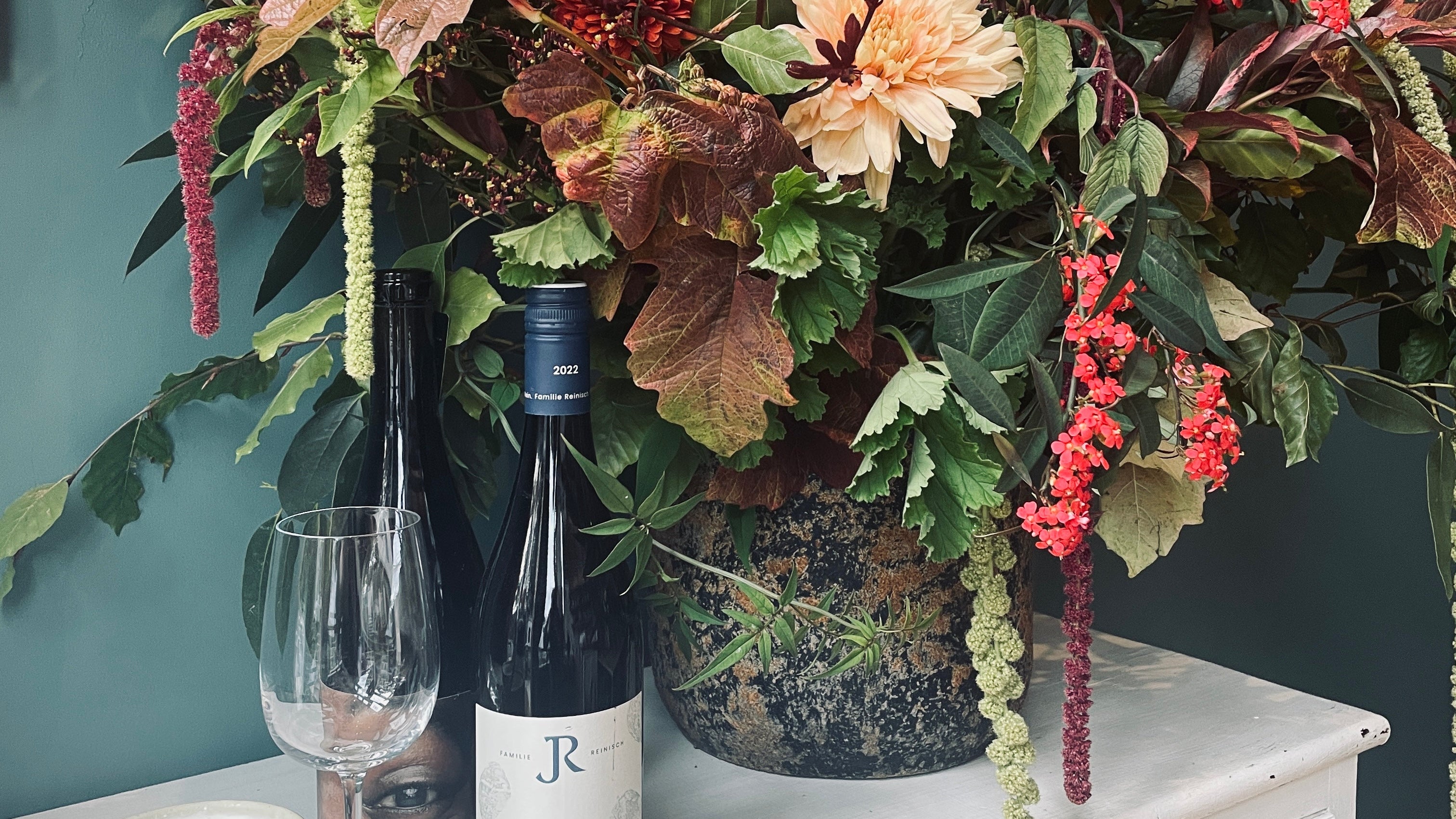 Floral arrangement with bottles and a candle on a white surface against a gray wall.