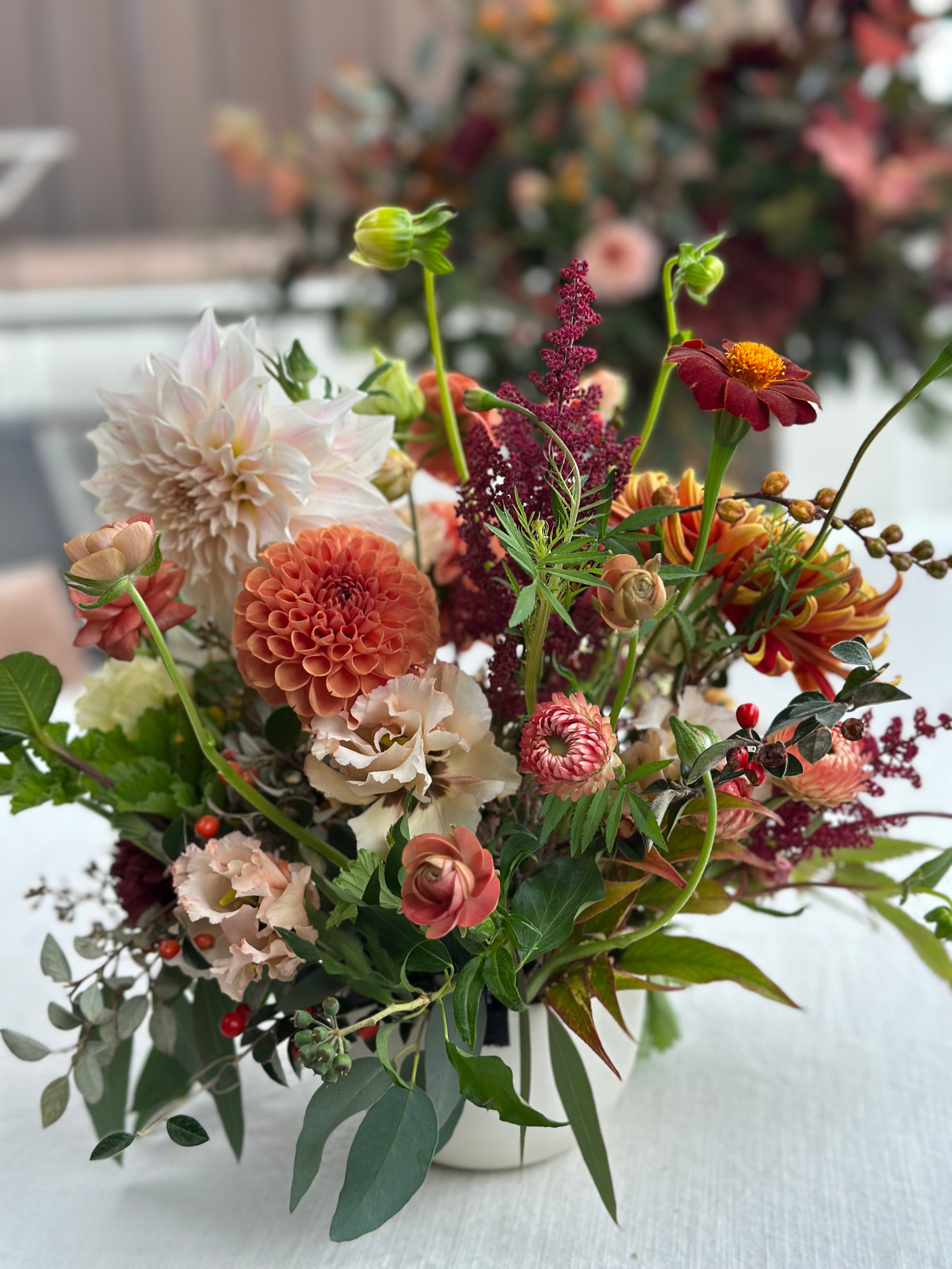 A bowl of flowers in Oranges, Creams and Burgundy's in the foreground, with another larger arrangement blurred in the background