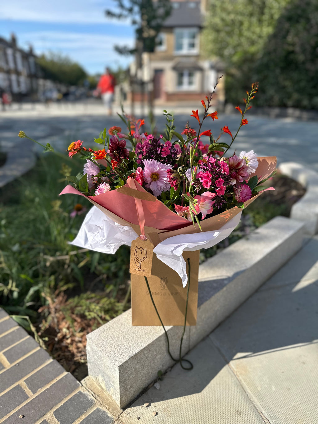 A hand-tied flower bouquet with pink, purple, and orange blossoms wrapped in pink and white tissue paper and a kraft paper bag, sitting on a stone curb outdoors.
