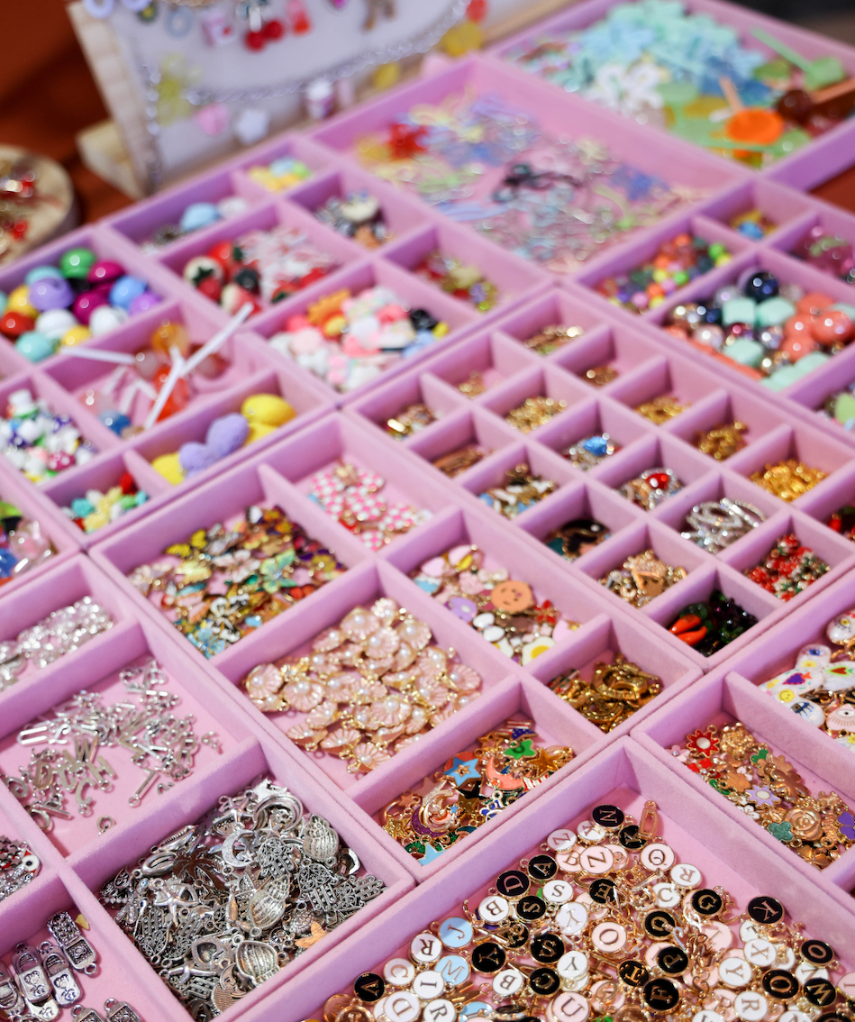 A pink organizer tray filled with many small compartments containing assorted craft embellishments, beads, buttons, rhinestones, and metallic charms in assorted colors.
