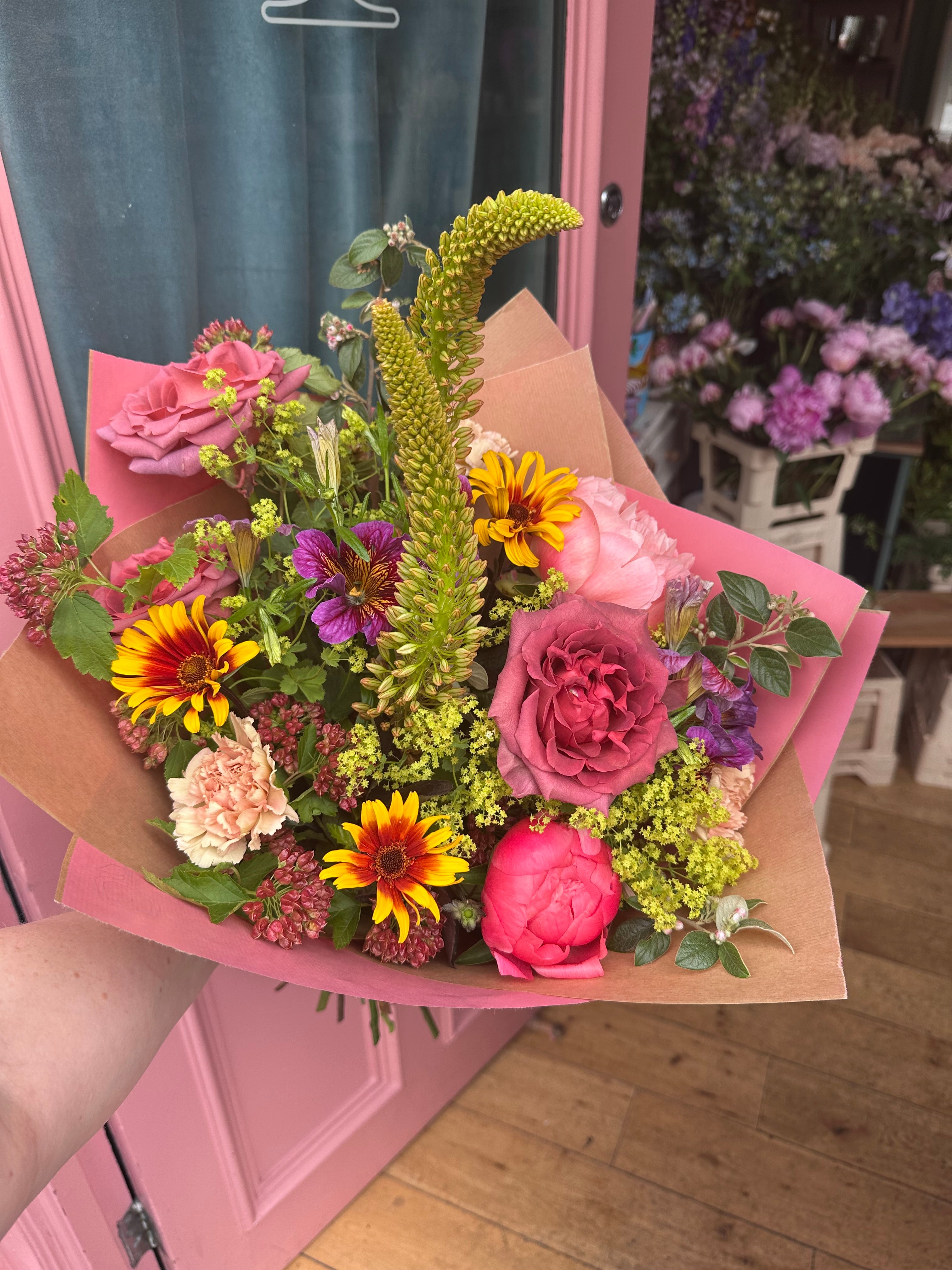 A hand holding a vibrant mixed flower bouquet wrapped in pink paper, set against a pink door and wooden floor background.