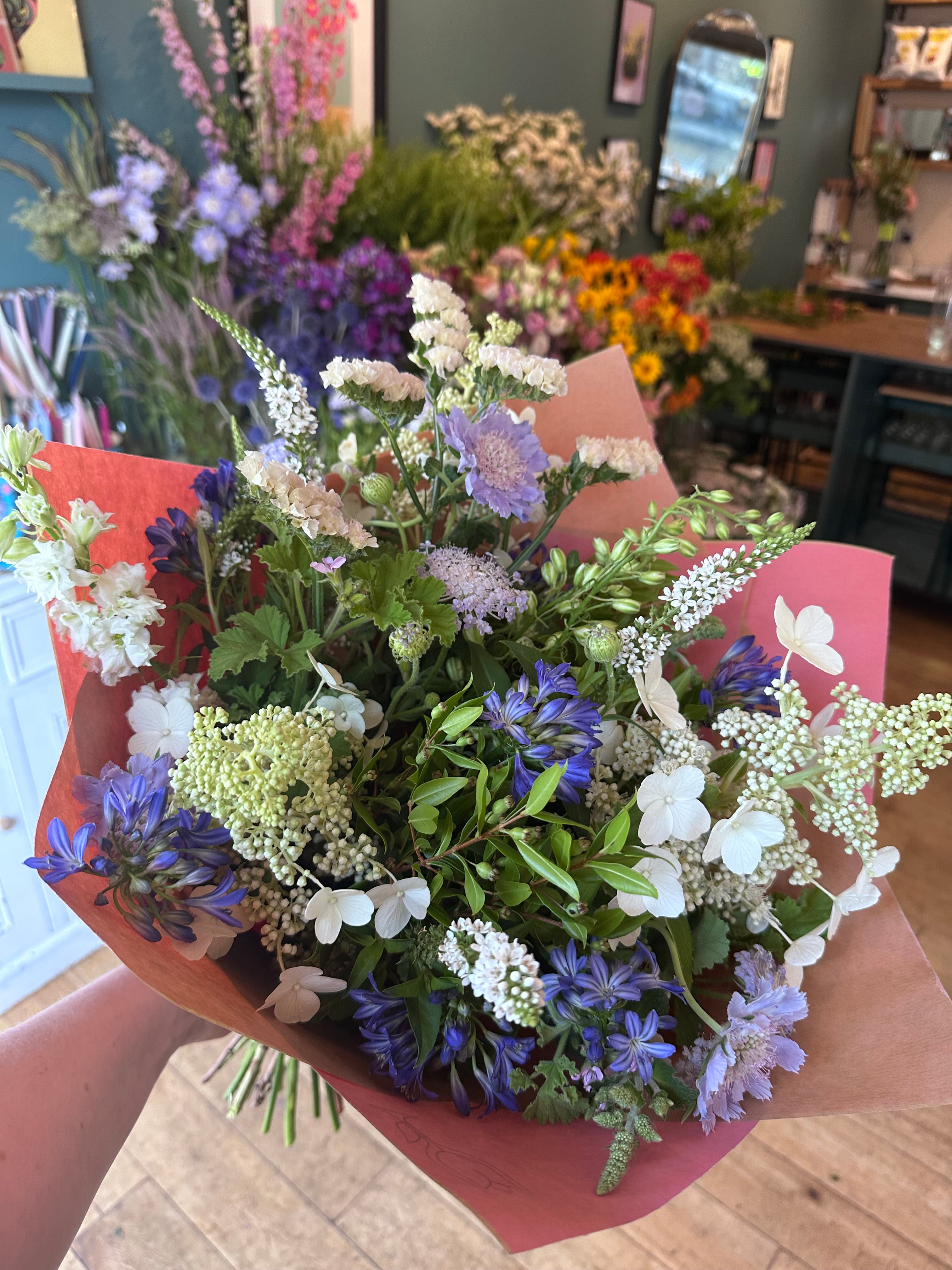 A hand holding a vibrant mixed flower bouquet wrapped in reddish-brown paper, set against a softly blurred background of a flower shop interior.